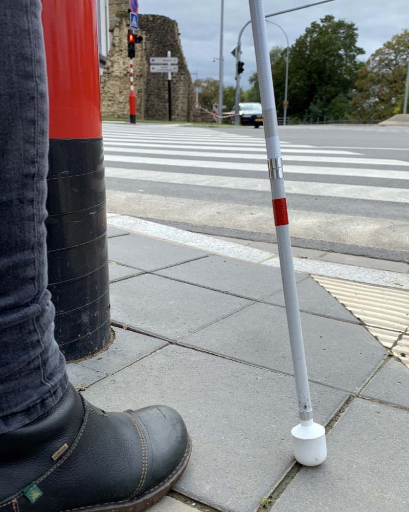 Blindenlangstock und Bein neben Ampelpfosten, im Hintergrund die Ampel der anderen Straßenseite, die auf rot steht.